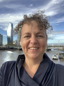 Portrait photo of Nicole Brown. White woman with curly, frizzy hair, blue eyes, wearing a blue top. The Thames and London Southbank are visible in the background.